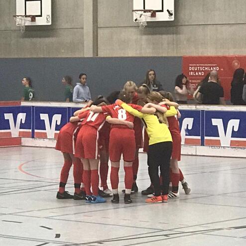 Mädchenmannschaft in roten Trikots umarmt sich zur Besprechung in einer Sporthalle vor einem Turnier.