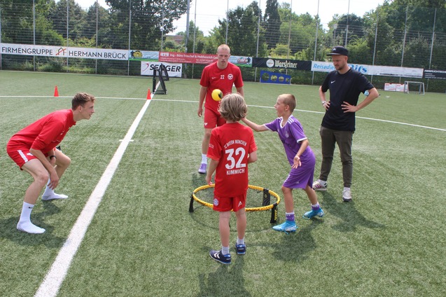 Kinder spielen mit einem Tennisball in einem Trainingsspiel, w&auml;hrend zwei Trainer zuschauen. Sportplatz im Hintergrund.