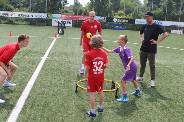Kinder spielen mit einem Tennisball in einem Trainingsspiel, w&auml;hrend zwei Trainer zuschauen. Sportplatz im Hintergrund.