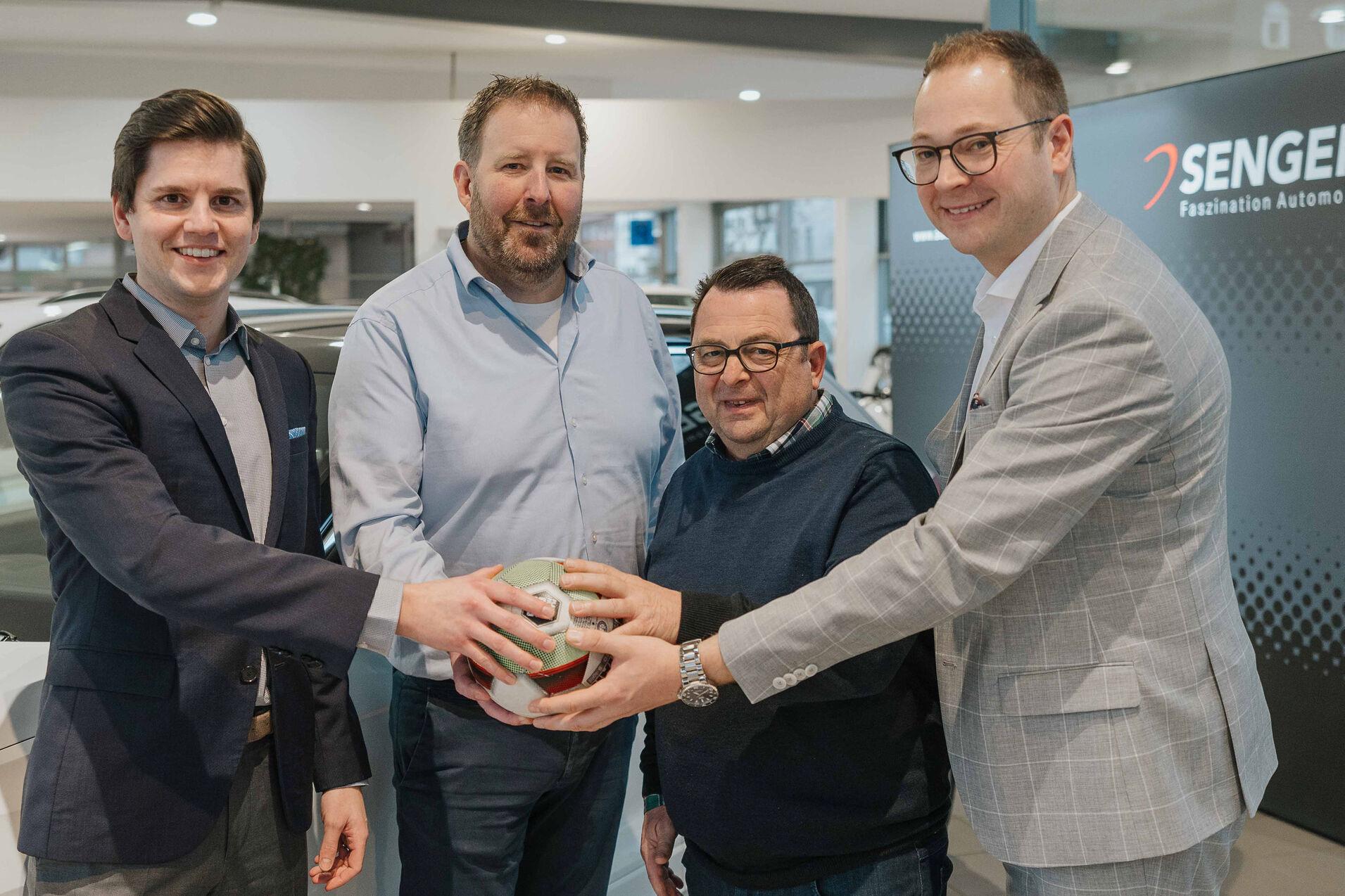 Vier Männer stehen in einem Autohaus und halten gemeinsam einen Fußball, im Hintergrund das SENGER-Logo sichtbar.