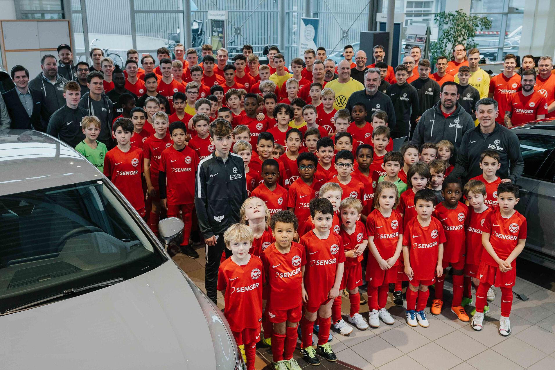 Gruppenfoto von Fu&szlig;ballspielern in roten Trikots, umgeben von Trainern und Erwachsenen in einem Autohaus.