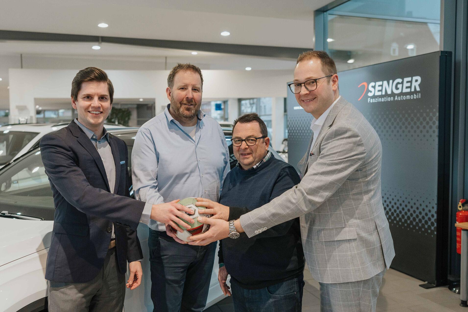 Vier M&auml;nner stehen in einem Autohaus und halten gemeinsam einen Fu&szlig;ball, im Hintergrund das SENGER-Logo sichtbar.
