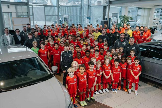 Gruppenfoto von Fu&szlig;ballspielern in roten Trikots, umgeben von Trainern und Erwachsenen in einem Autohaus.