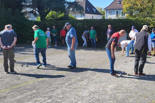 Mehrere Personen spielen Boule auf einem Platz, umgeben von Zuschauern und B&auml;umen im Hintergrund.
