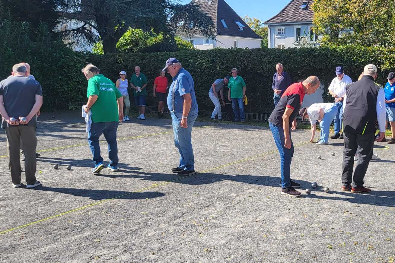 Mehrere Personen spielen Boule auf einem Platz, umgeben von Zuschauern und B&auml;umen im Hintergrund.