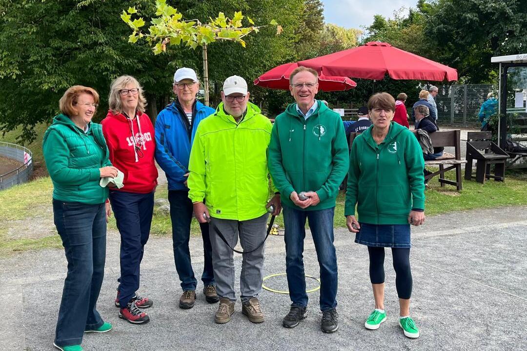 Sechs Personen in gr&uuml;nen Jacken stehen auf einem Platz mit B&auml;umen und einem Kaffeestand im Hintergrund.