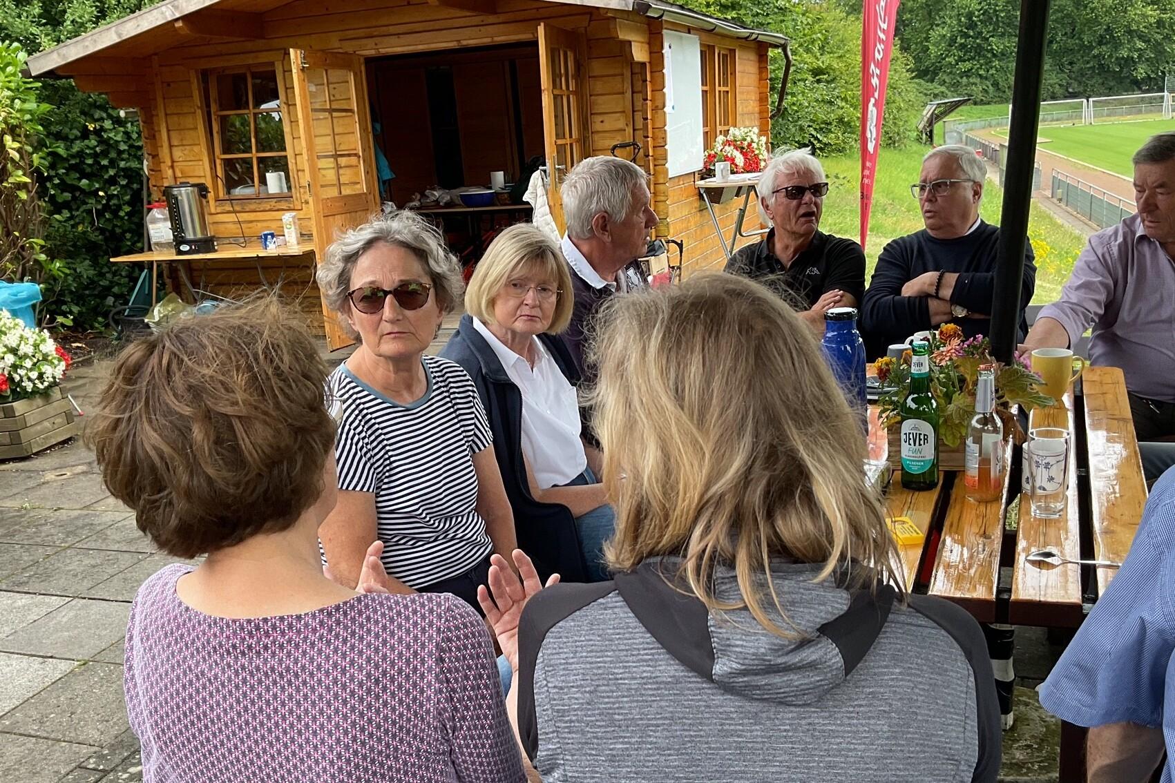 Gruppe von Menschen sitzt an einem Tisch im Freien, spricht und genie&szlig;t Getr&auml;nke vor einer Holzh&uuml;tte.