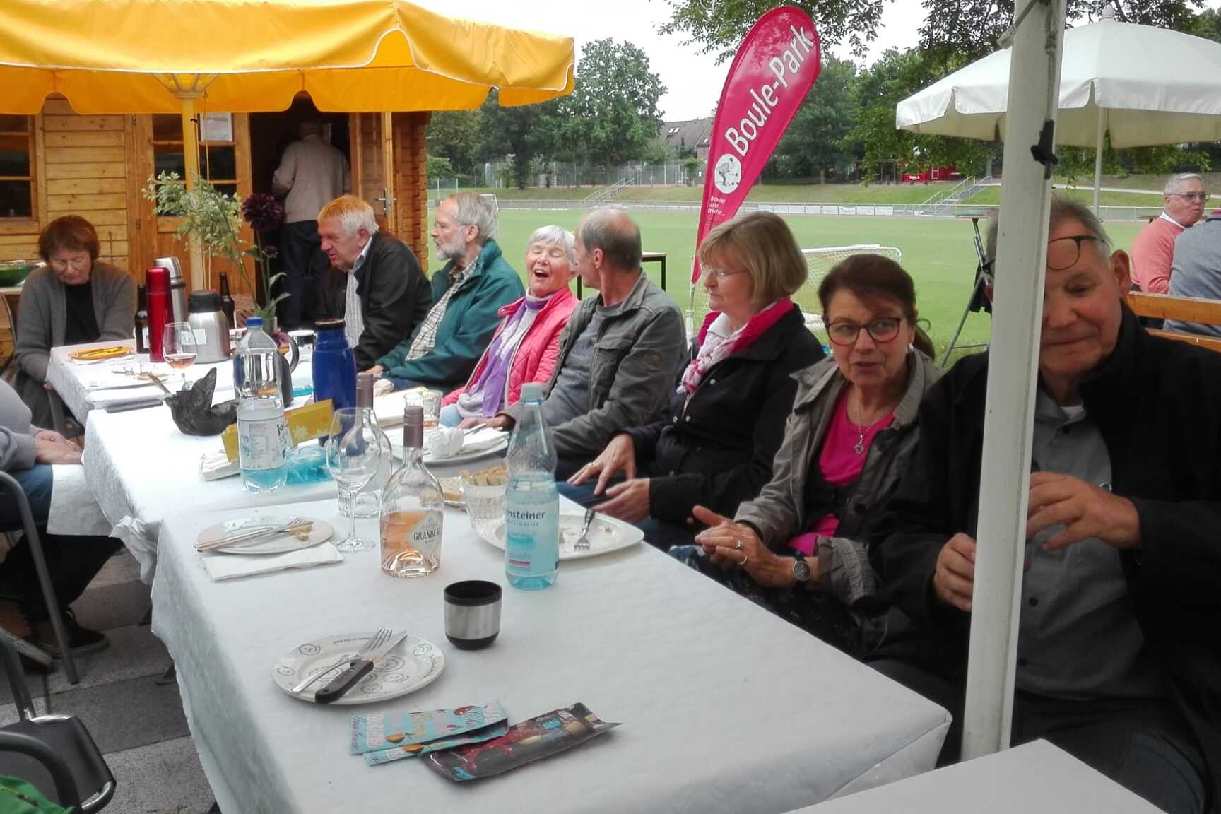 Gruppenszene mit mehreren Personen an einem gedeckten Tisch unter Sonnenschirmen in einem Freiluftbereich.