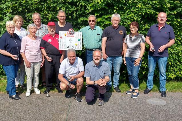 Gruppe von elf Personen posiert l&auml;chelnd vor einer gr&uuml;nen Hecke, einige halten Plakate oder Becher in der Hand.