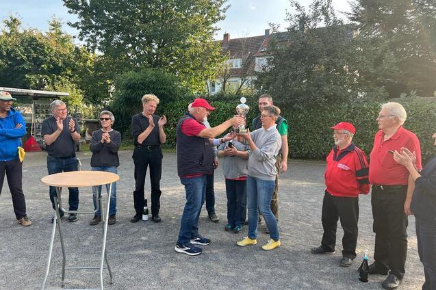 Siegerehrung mit mehreren Personen, die einem Gewinner einen Pokal &uuml;berreichen, umringt von applaudierenden Zuschauern.