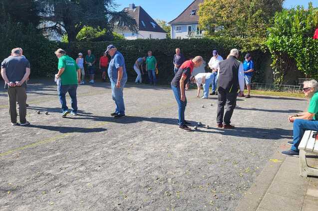 Mehrere Personen spielen Boule auf einem Platz, umgeben von Zuschauern und B&auml;umen im Hintergrund.