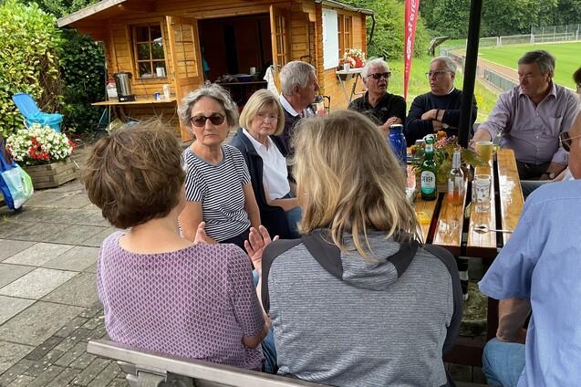 Gruppe von Menschen sitzt an einem Tisch im Freien, spricht und genie&szlig;t Getr&auml;nke vor einer Holzh&uuml;tte.