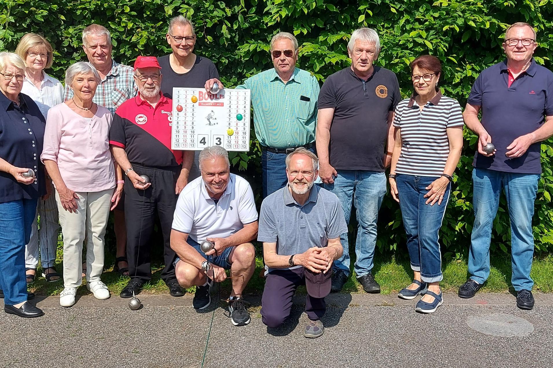 Gruppe von elf Personen posiert l&auml;chelnd vor einer gr&uuml;nen Hecke, einige halten Plakate oder Becher in der Hand.