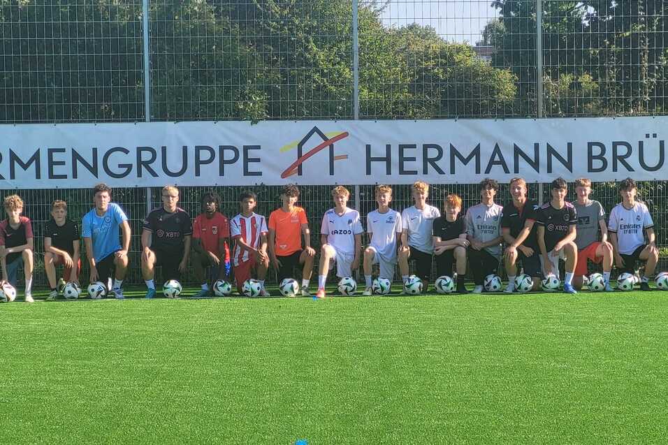 Gruppe von Jugendlichen mit Fu&szlig;ballschuhen, die auf einem Kunstrasenplatz vor einem Werbebanner posieren.
