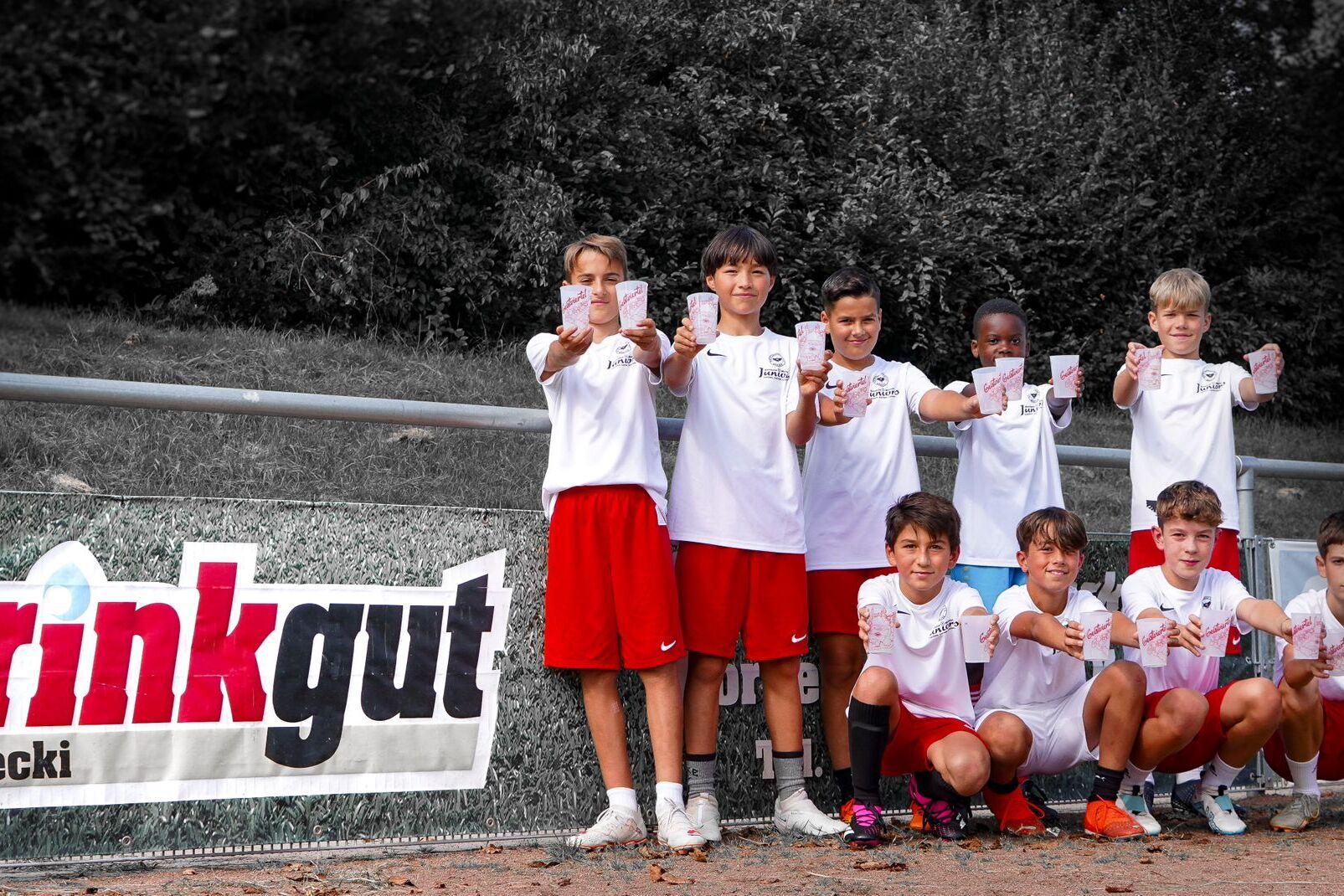 Gruppe von sieben Kindern in Sportkleidung, die stolz im Vordergrund stehen und Zettel in die Kamerarichtung halten, vor Banner.