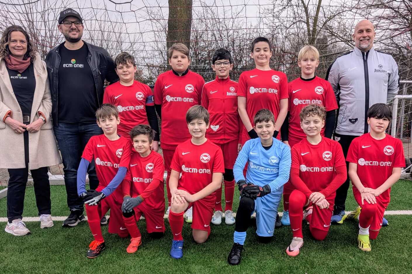 Gruppensfoto einer Jugendfußballmannschaft in roten Trikots mit Betreuern auf einem Sportplatz.