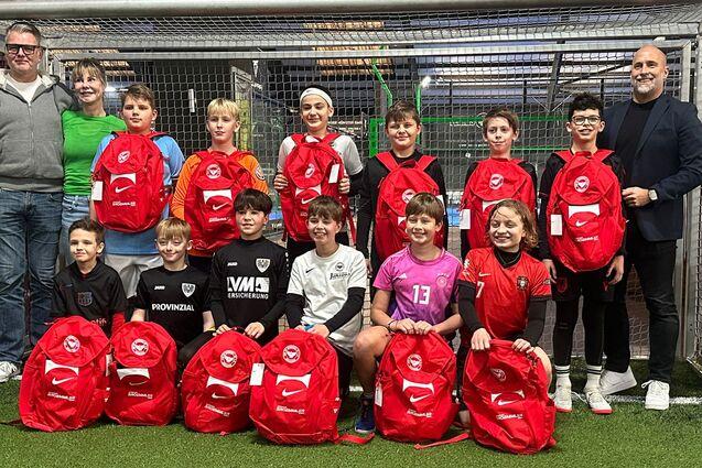 Gruppe von Kindern und Erwachsenen in einer Sporthalle, posierend vor einem Tor mit roten Rucks&auml;cken.