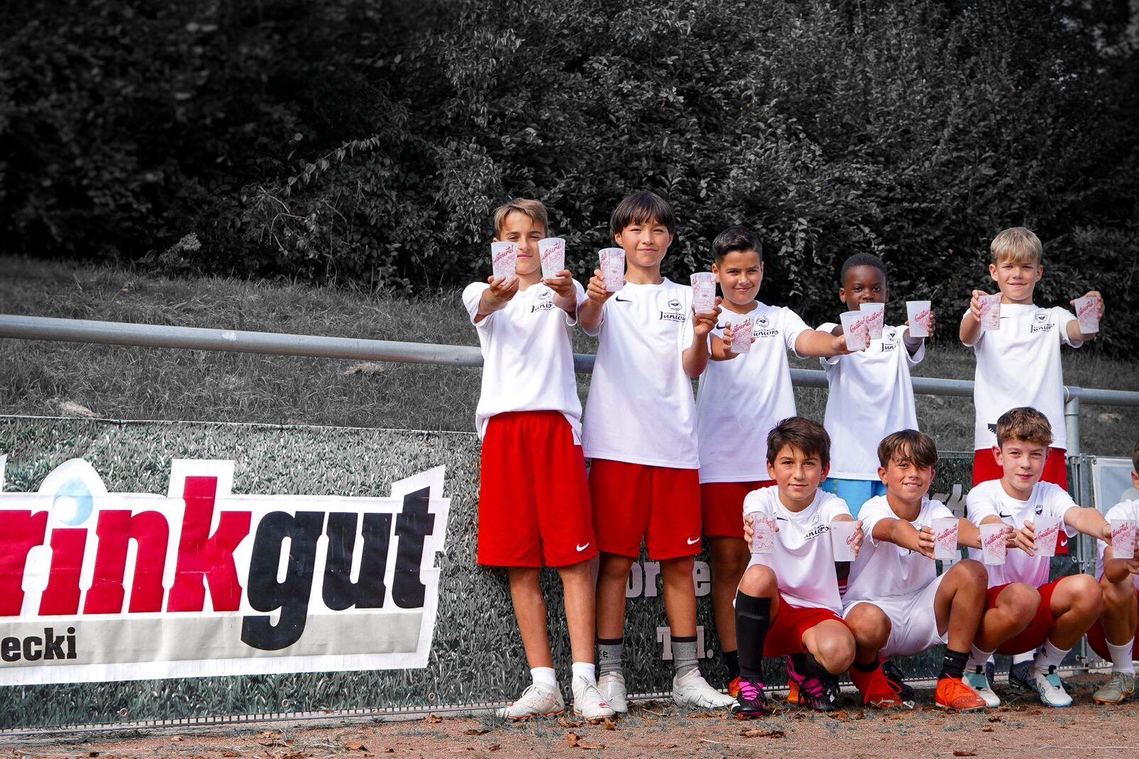Gruppe von sieben Kindern in Sportkleidung, die stolz im Vordergrund stehen und Zettel in die Kamerarichtung halten, vor Banner.