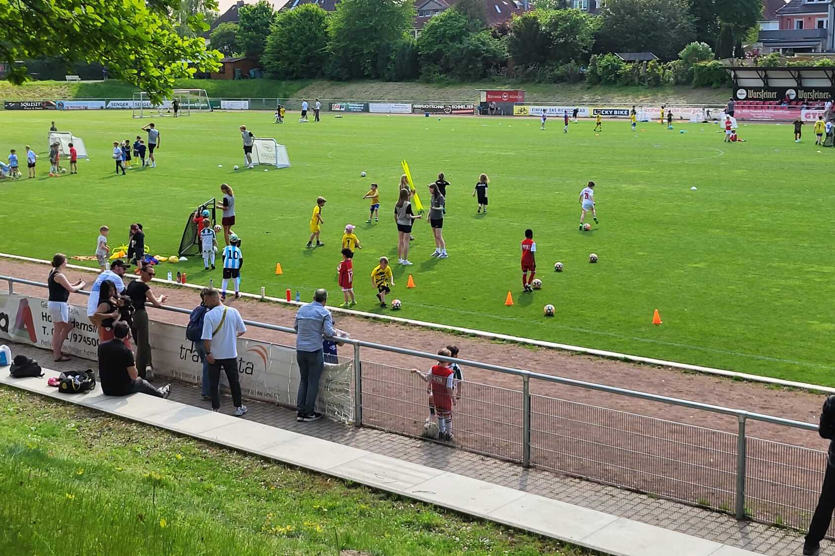 Kindermannschaften trainieren auf einem gr&uuml;nen Fu&szlig;ballplatz, umgeben von Zuschauern und weiteren Aktivit&auml;ten.