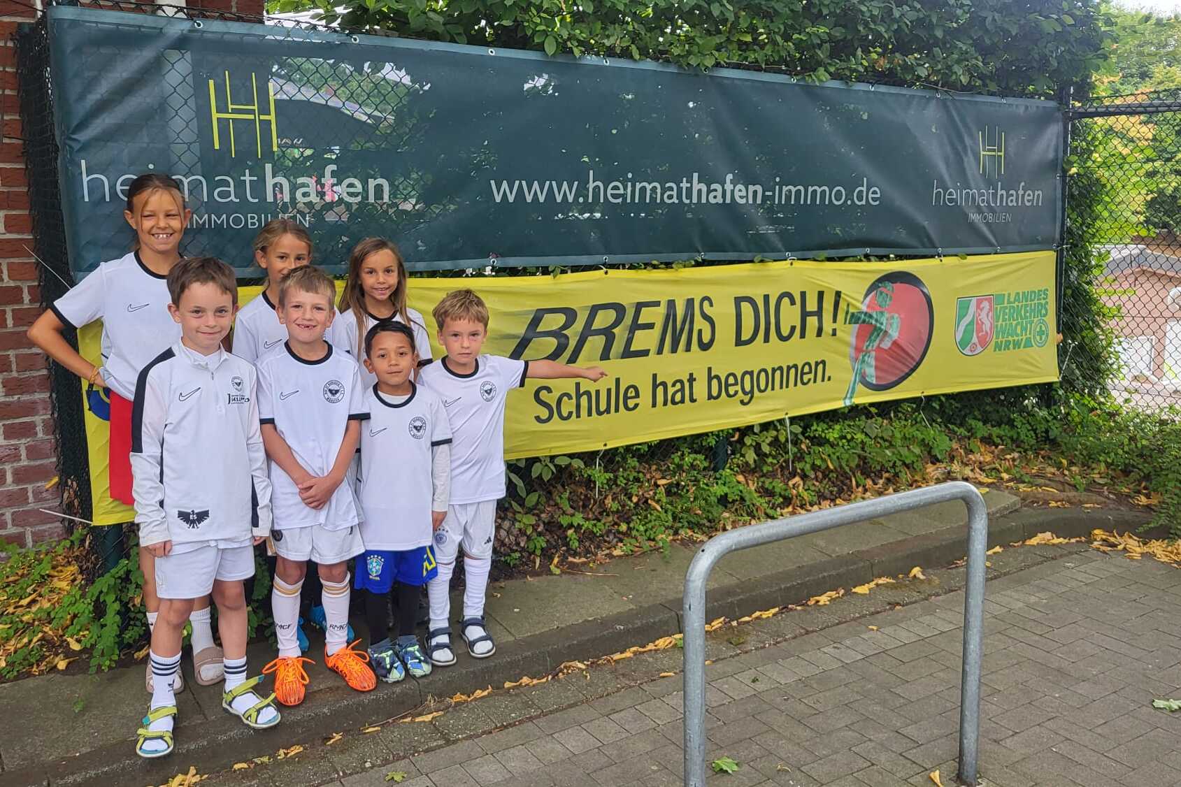 Gruppe von Kindern in Fu&szlig;balltrikots steht l&auml;chelnd vor einem Banner mit der Aufschrift &bdquo;BREMS DICH! Schule hat begonnen.&ldquo;
