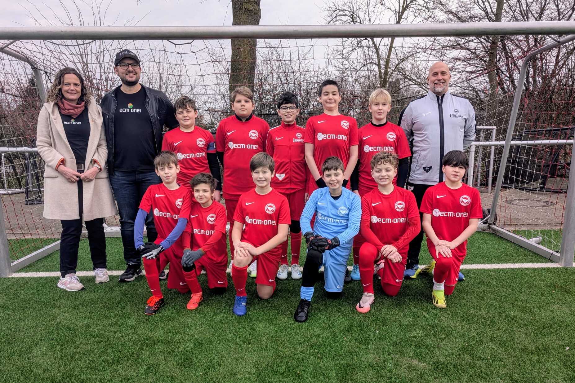 Gruppensfoto einer Jugendfu&szlig;ballmannschaft in roten Trikots mit Betreuern auf einem Sportplatz.