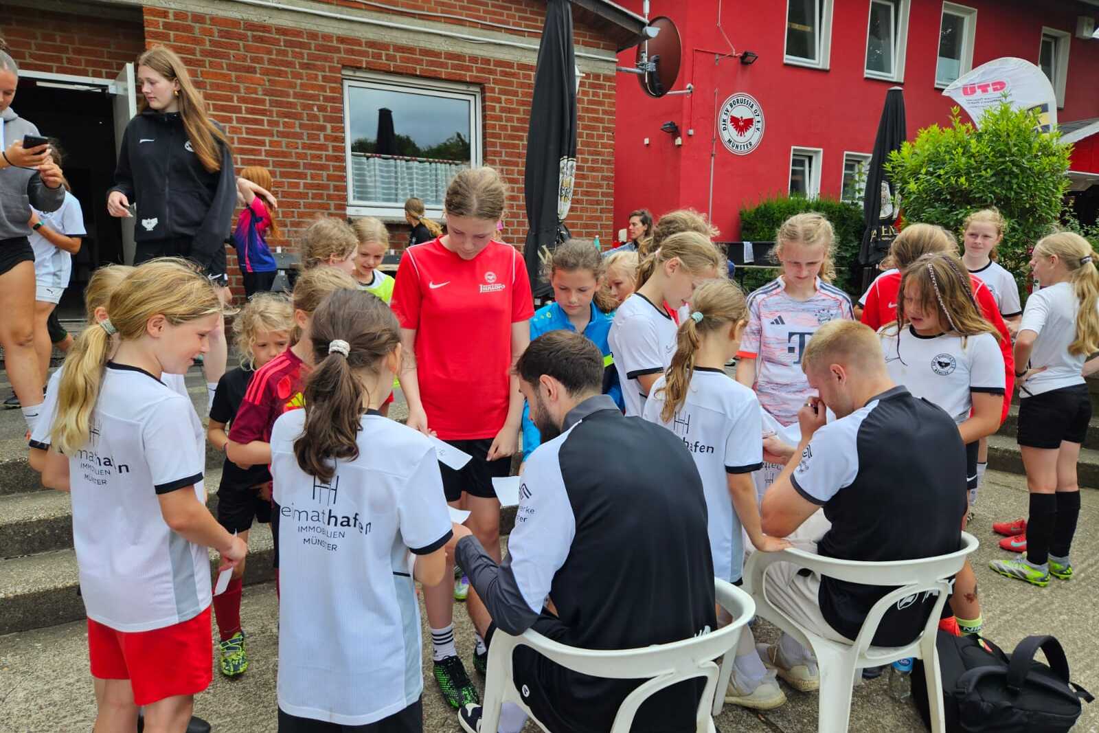 Gruppe von M&auml;dchen in Fu&szlig;balltrikots umringt Trainer, der Autogramme auf einem Platz vor einem Geb&auml;ude gibt.