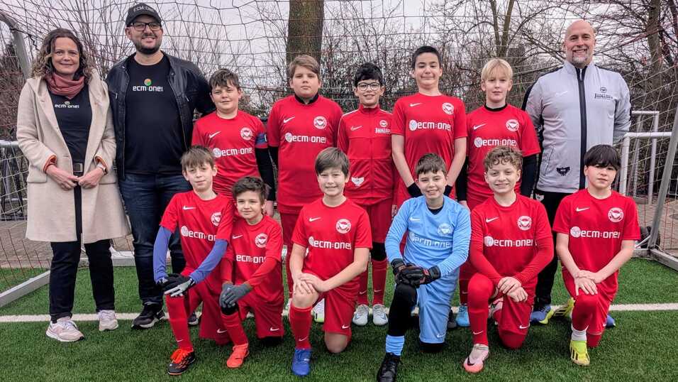 Gruppensfoto einer Jugendfu&szlig;ballmannschaft in roten Trikots mit Betreuern auf einem Sportplatz.