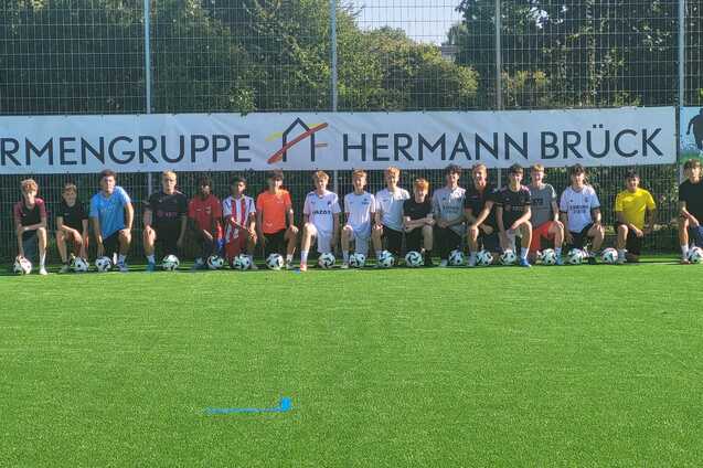 Gruppe von Jugendlichen mit Fu&szlig;ballschuhen, die auf einem Kunstrasenplatz vor einem Werbebanner posieren.
