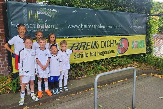 Gruppe von Kindern in Fu&szlig;balltrikots steht l&auml;chelnd vor einem Banner mit der Aufschrift &bdquo;BREMS DICH! Schule hat begonnen.&ldquo;