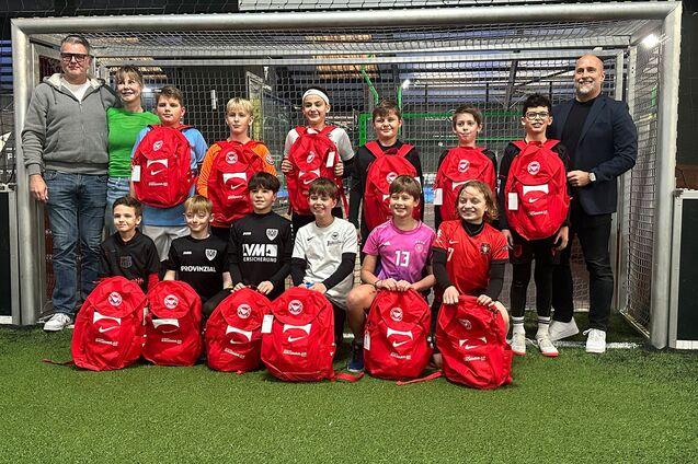 Gruppe von Kindern und Erwachsenen in einer Sporthalle, posierend vor einem Tor mit roten Rucks&auml;cken.