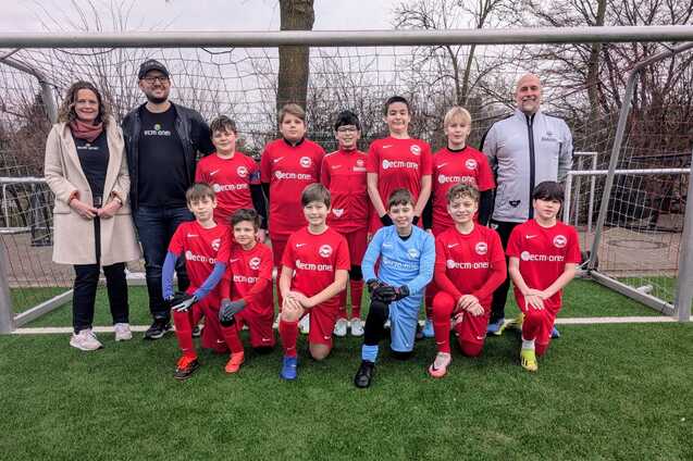 Gruppensfoto einer Jugendfu&szlig;ballmannschaft in roten Trikots mit Betreuern auf einem Sportplatz.