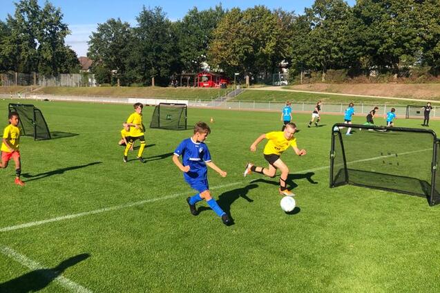 Zwei Kinder in Fu&szlig;balltrikots rennen auf einem Rasenplatz auf einen Ball zu, mit Toren im Hintergrund.