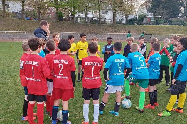Gruppe von Jugendlichen in Fu&szlig;balltrikots versammelt sich um einen Trainer auf einem Rasenplatz.