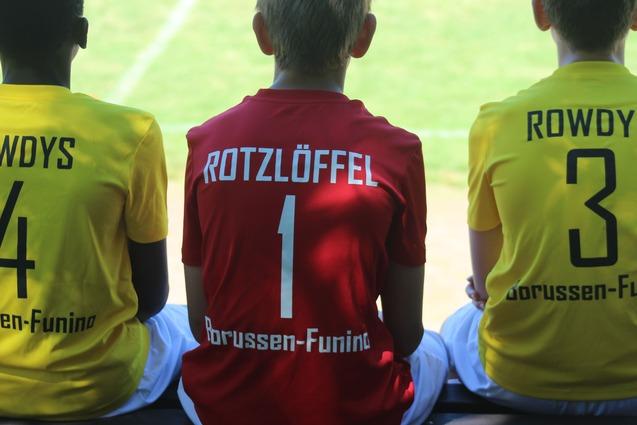 Drei Kinder in Trikots sitzen nebeneinander auf einer Bank, mit den R&uuml;ckennummern 4, 1 und 3.