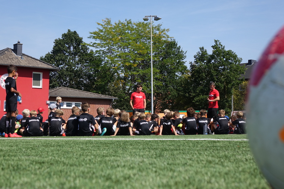 Gruppe von Kindern in schwarzen Trikots sitzt auf einem Fu&szlig;ballplatz, w&auml;hrend Trainer Anweisungen gibt. Ein Fu&szlig;ball im Vordergrund.