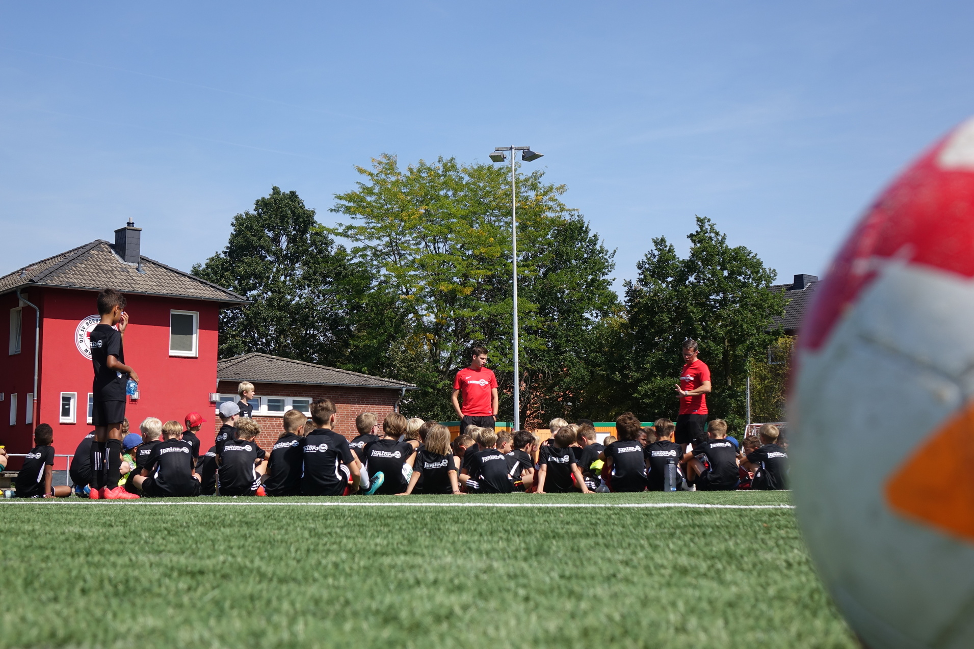 Gruppe von Kindern in schwarzen Trikots sitzt auf einem Fu&szlig;ballplatz, w&auml;hrend Trainer Anweisungen gibt. Ein Fu&szlig;ball im Vordergrund.