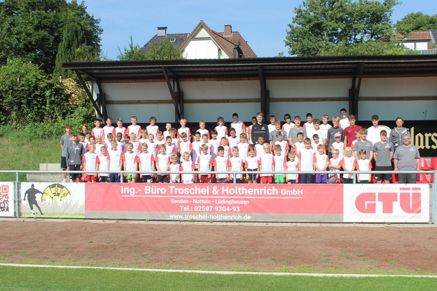 Gruppenfoto von Spielern und Trainern auf einer Sportanlage, alle tragen einheitliche Trikots und stehen auf einer Trib&uuml;ne.