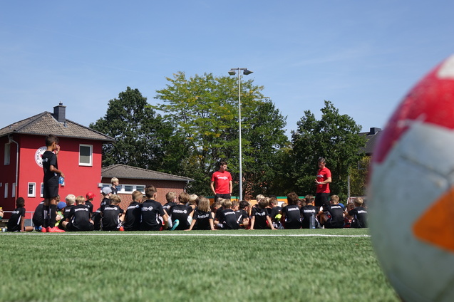 Gruppe von Kindern in schwarzen Trikots sitzt auf einem Fu&szlig;ballplatz, w&auml;hrend Trainer Anweisungen gibt. Ein Fu&szlig;ball im Vordergrund.
