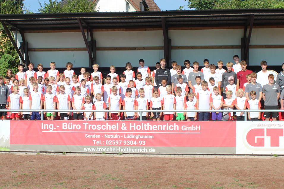 Gruppenfoto von Spielern und Trainern auf einer Sportanlage, alle tragen einheitliche Trikots und stehen auf einer Trib&uuml;ne.