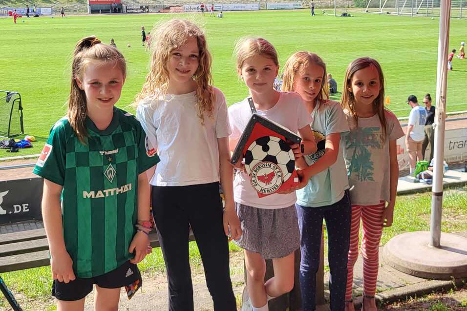 F&uuml;nf M&auml;dchen stehen l&auml;chelnd auf einem Sportplatz, halten ein Fu&szlig;ball-Troph&auml;e und tragen sportliche Kleidung.