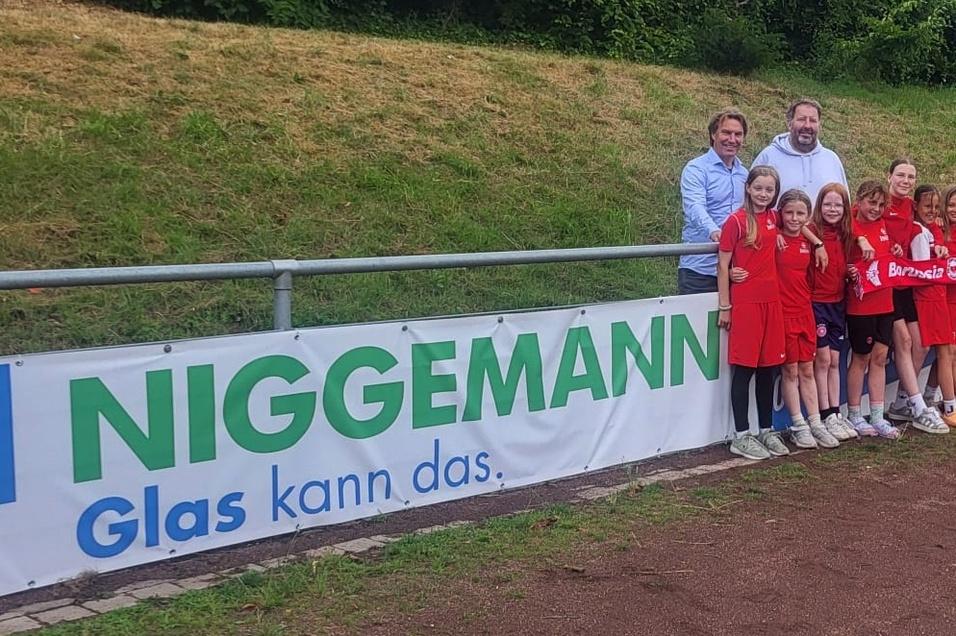 Gruppenshot von zwei Erwachsenen und neun M&auml;dchen in roten Trikots vor einem Sponsorenbanner am Fu&szlig;ballplatz.