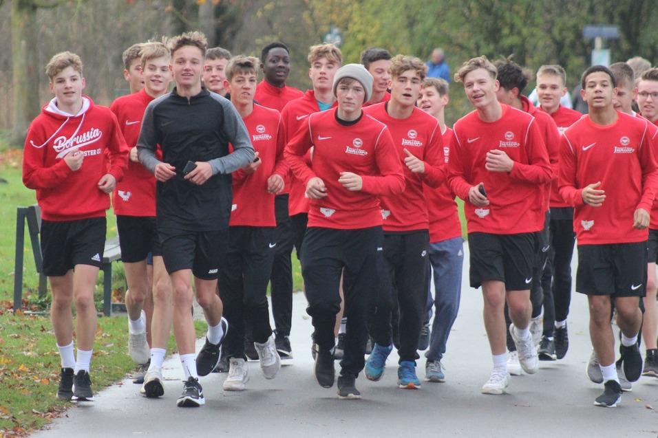 Gruppe von Jugendlichen in roten Trainingsshirts joggt auf einem Weg durch einen Park im Herbst.