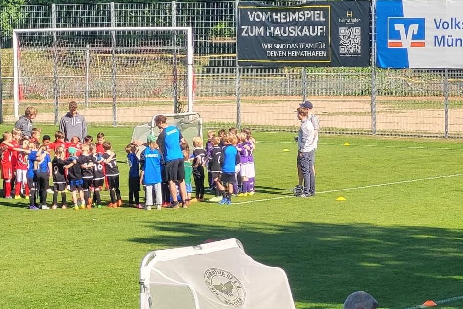 Gruppierung von Kindern in Sportkleidung auf einem Fu&szlig;ballplatz, umrahmt von Trainern und einer Werbebehinderung.