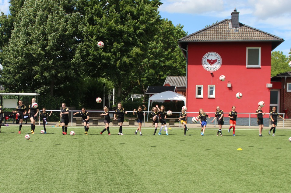 Gruppe von Fu&szlig;ballspielern in Training, die B&auml;lle auf einem Kunstrasenplatz passen, im Hintergrund ein rotes Vereinsgeb&auml;ude.