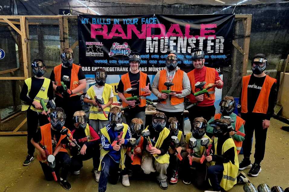 Gruppenselfie von Teilnehmern in Schutzkleidung mit Paintball-Ausr&uuml;stung vor einem Banner in einer Indoor-Spielarena.