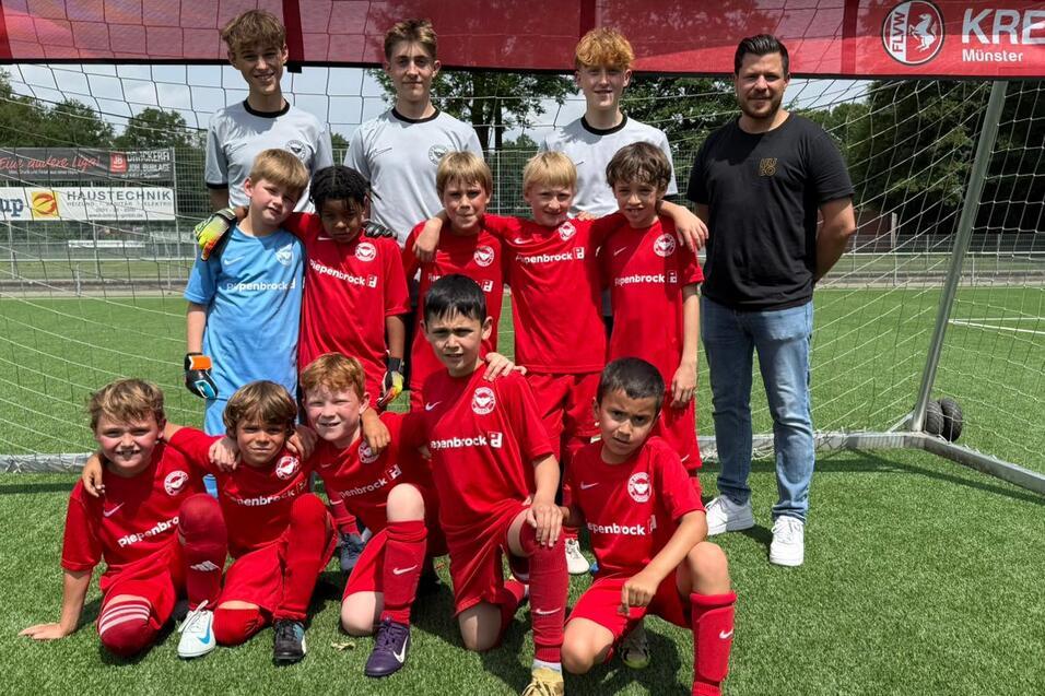 Gruppe von 13 jungen Fu&szlig;ballspielern in roten Trikots und zwei Trainern vor einem Tor auf einem Kunstrasenplatz.