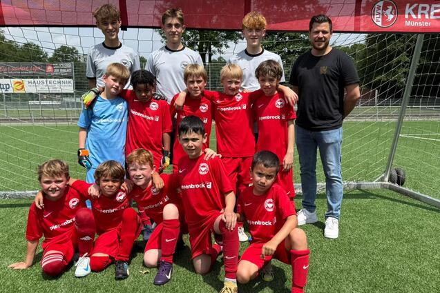 Gruppe von 13 jungen Fu&szlig;ballspielern in roten Trikots und zwei Trainern vor einem Tor auf einem Kunstrasenplatz.