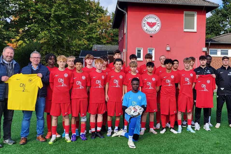 Jugendfu&szlig;ballmannschaft in roten Trikots steht vor einem Vereinsgeb&auml;ude, Trainer und Betreuer sind ebenfalls dabei.
