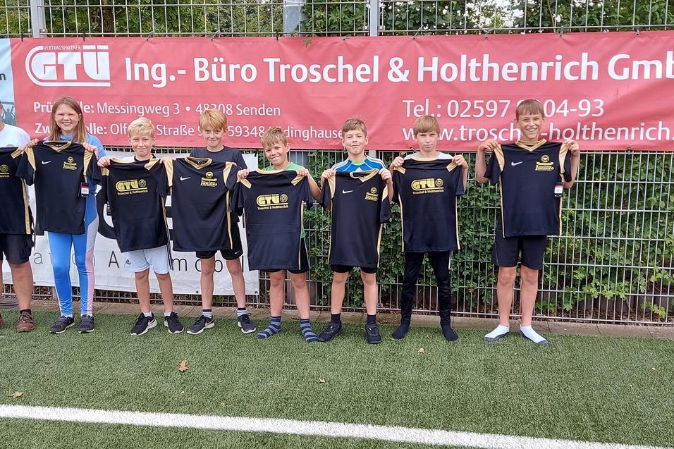 Gruppe von sieben Kindern in Sportbekleidung und einem Erwachsenen stehen vor einem Werbebanner auf einem Fu&szlig;ballfeld.