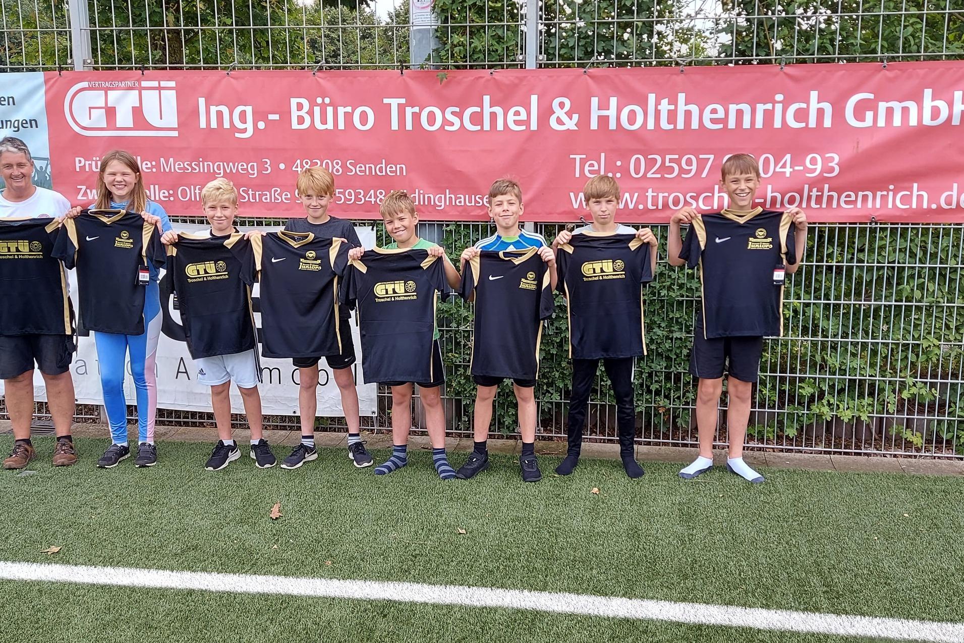 Gruppe von sieben Kindern in Sportbekleidung und einem Erwachsenen stehen vor einem Werbebanner auf einem Fu&szlig;ballfeld.